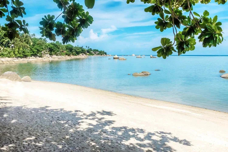 Tropical beach with white sand, calm blue water, green trees, and a partly cloudy sky.