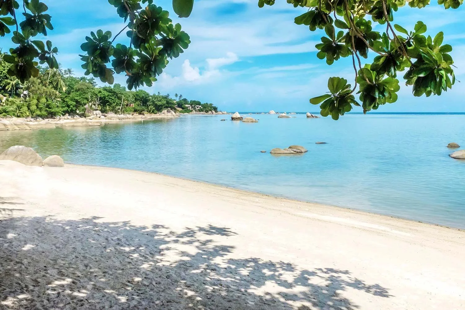 Tropical beach with white sand, calm blue water, green trees, and a partly cloudy sky.
